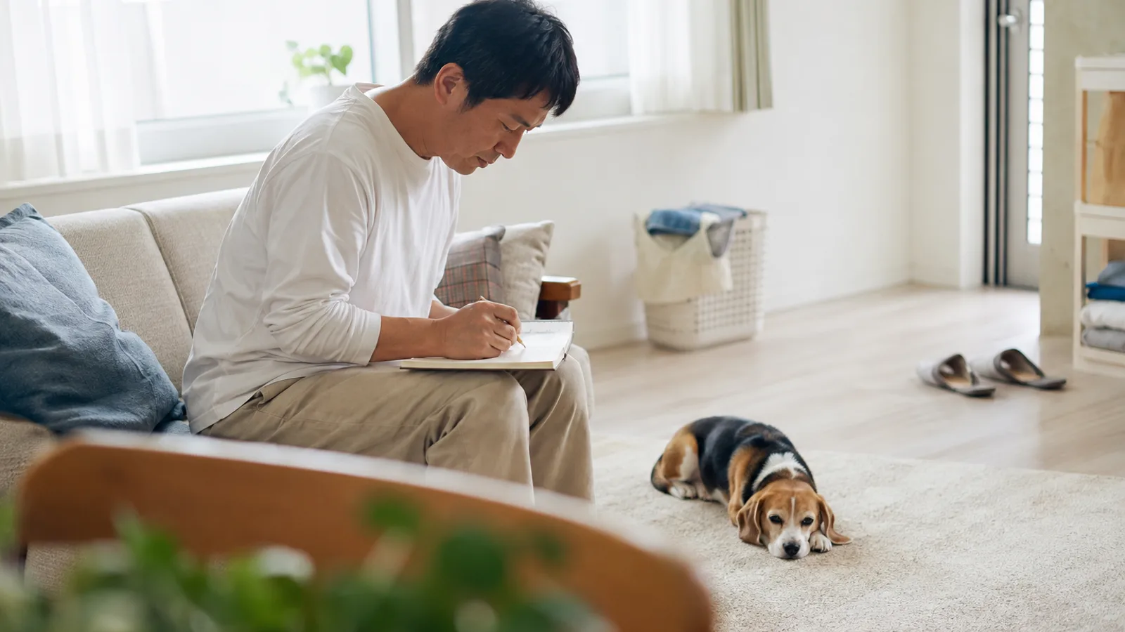 ソファでひざに広げた無地のノートに鉛筆で書き込む男性と、足元のラグで前足に顎をのせて伏せるトライカラーのビーグル