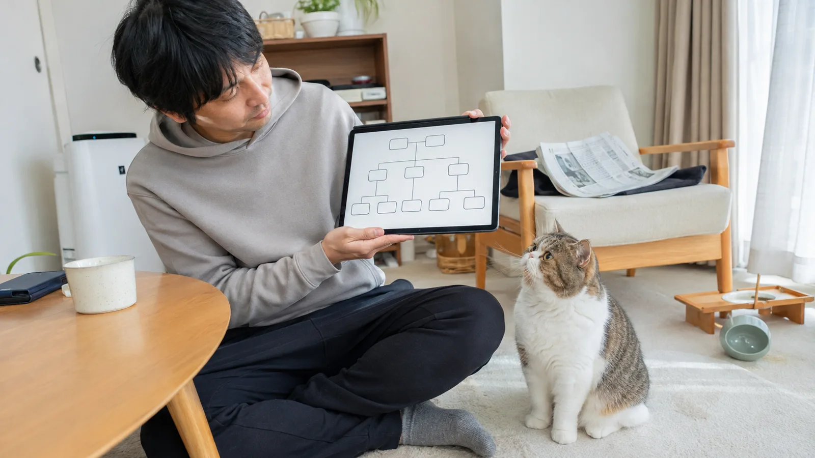 床でタブレットの家系図の画面を見せる男性と、そばのラグに座って画面を見上げる猫