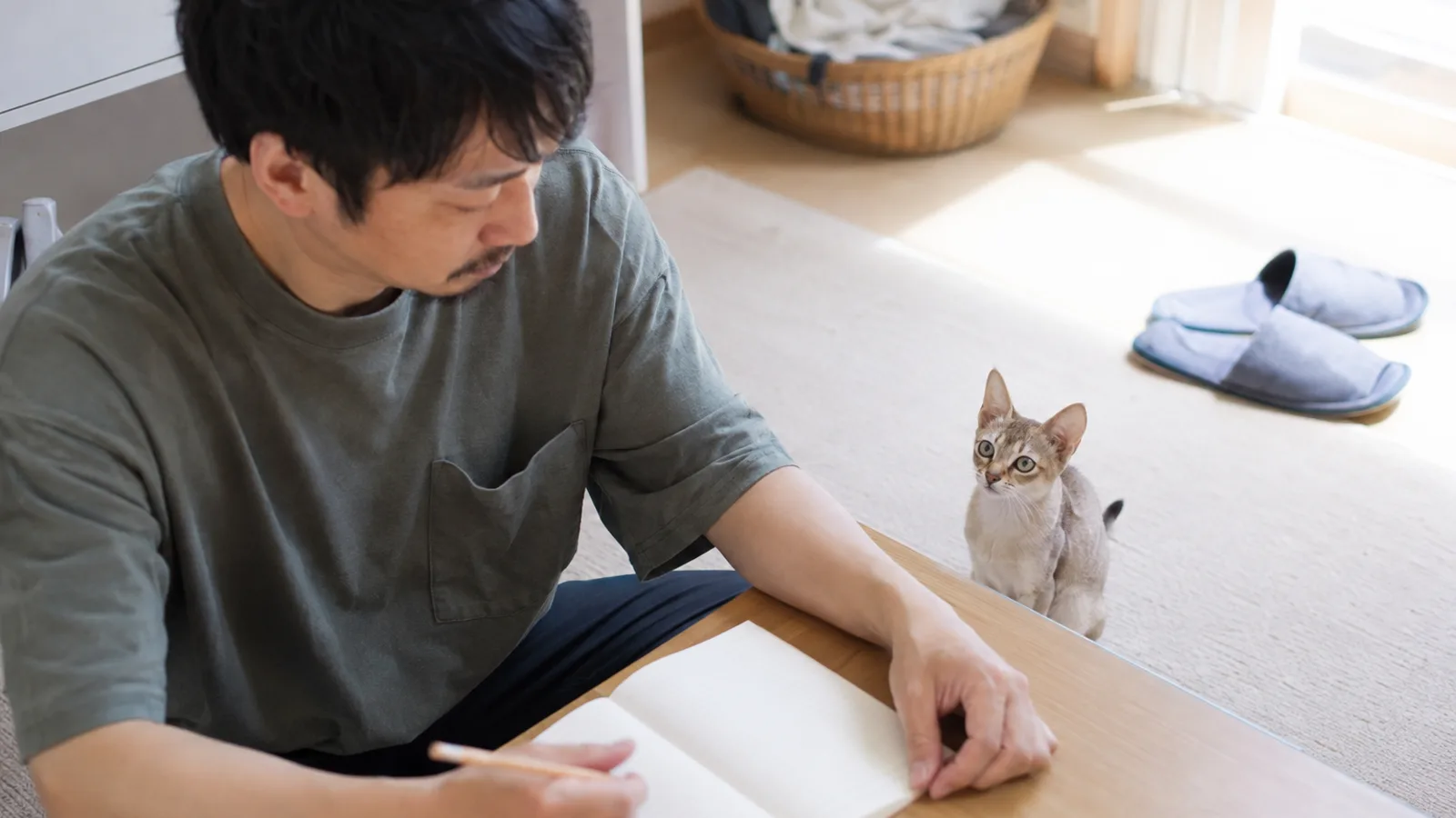 低いテーブルに開いた無地のノートに鉛筆で書き込む男性と、テーブルのそばの床に座って見上げるシンガプーラ