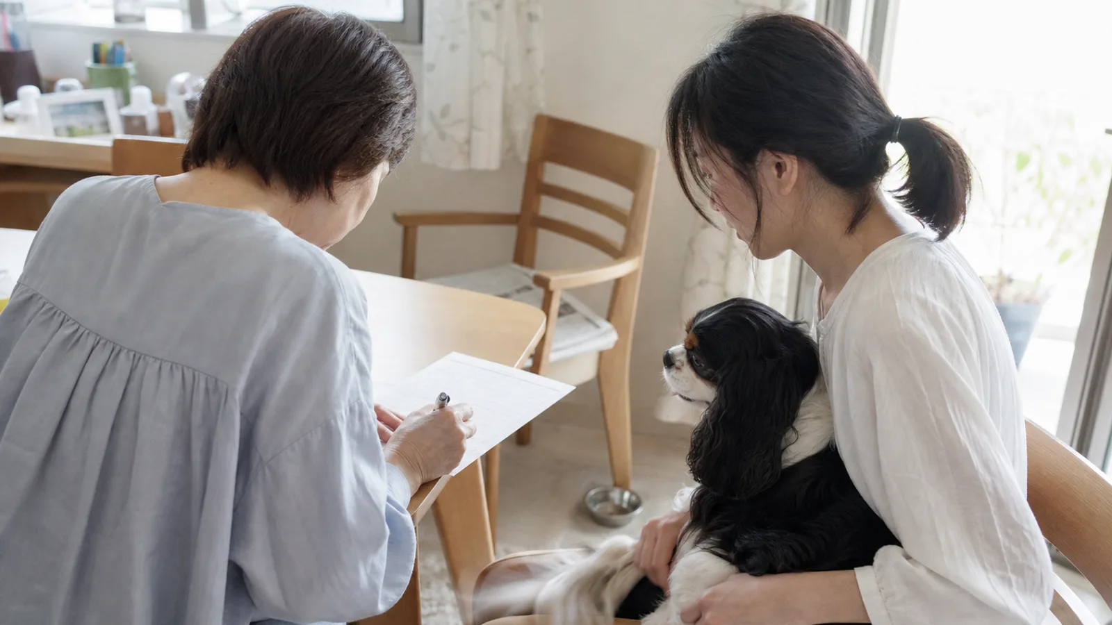 テーブルで書類に署名する50代の女性と、キャバリアを膝に抱いて見守る30代の女性