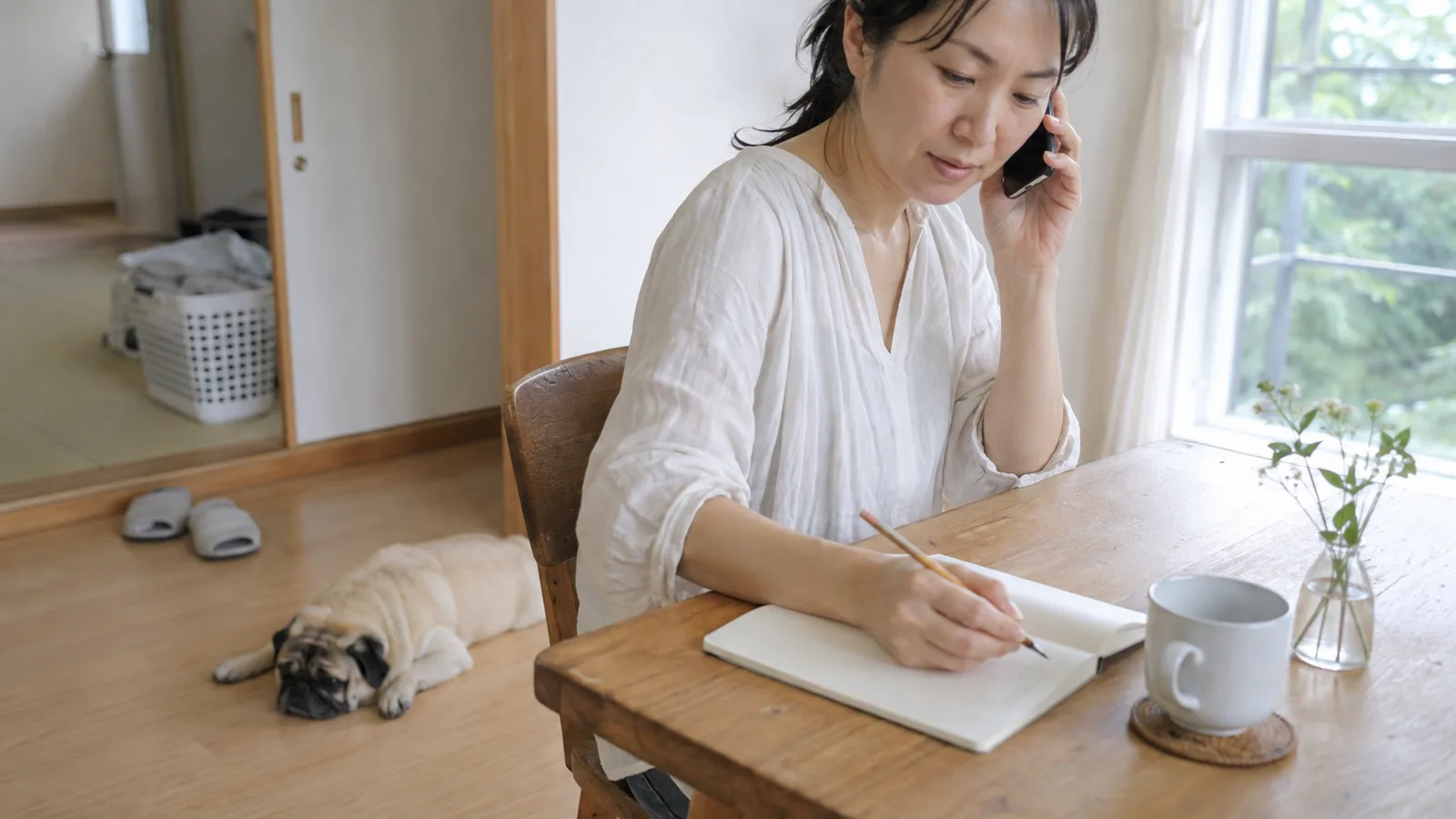 スマートフォンを耳に当てて話しながら無地のノートに書き留める女性と、足元で伏せるフォーンのパグ