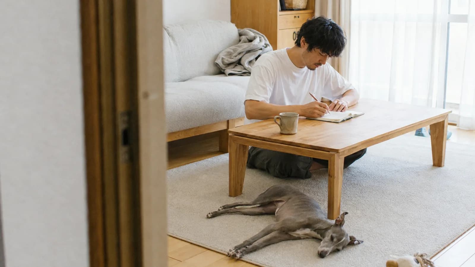 ローテーブルの小さな手帳に鉛筆で書き込む男性と、手前のラグに横向きに伏せるグレーのイタリアン・グレーハウンド