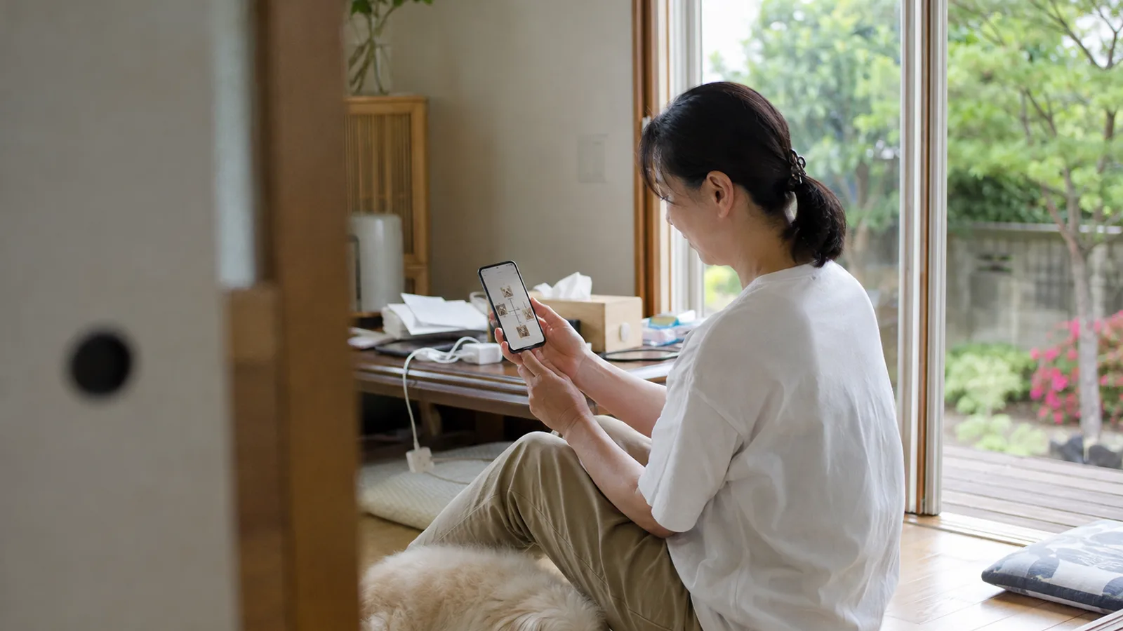 開け放した窓ぎわに座ってスマホの家系図画面を見る女性と、そのそばに寝そべるクリーム色の長い毛の犬