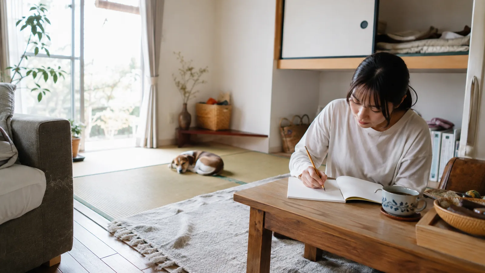 ちゃぶ台のノートに鉛筆で書き込む女性と、少し離れた畳の上で丸くなって眠る短毛の子犬