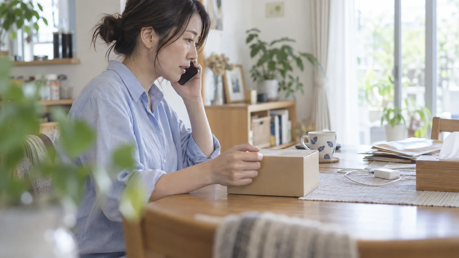 明るいダイニングのテーブルで、小さな無地の箱に片手を添え、スマートフォンを耳に当てて電話をかける女性