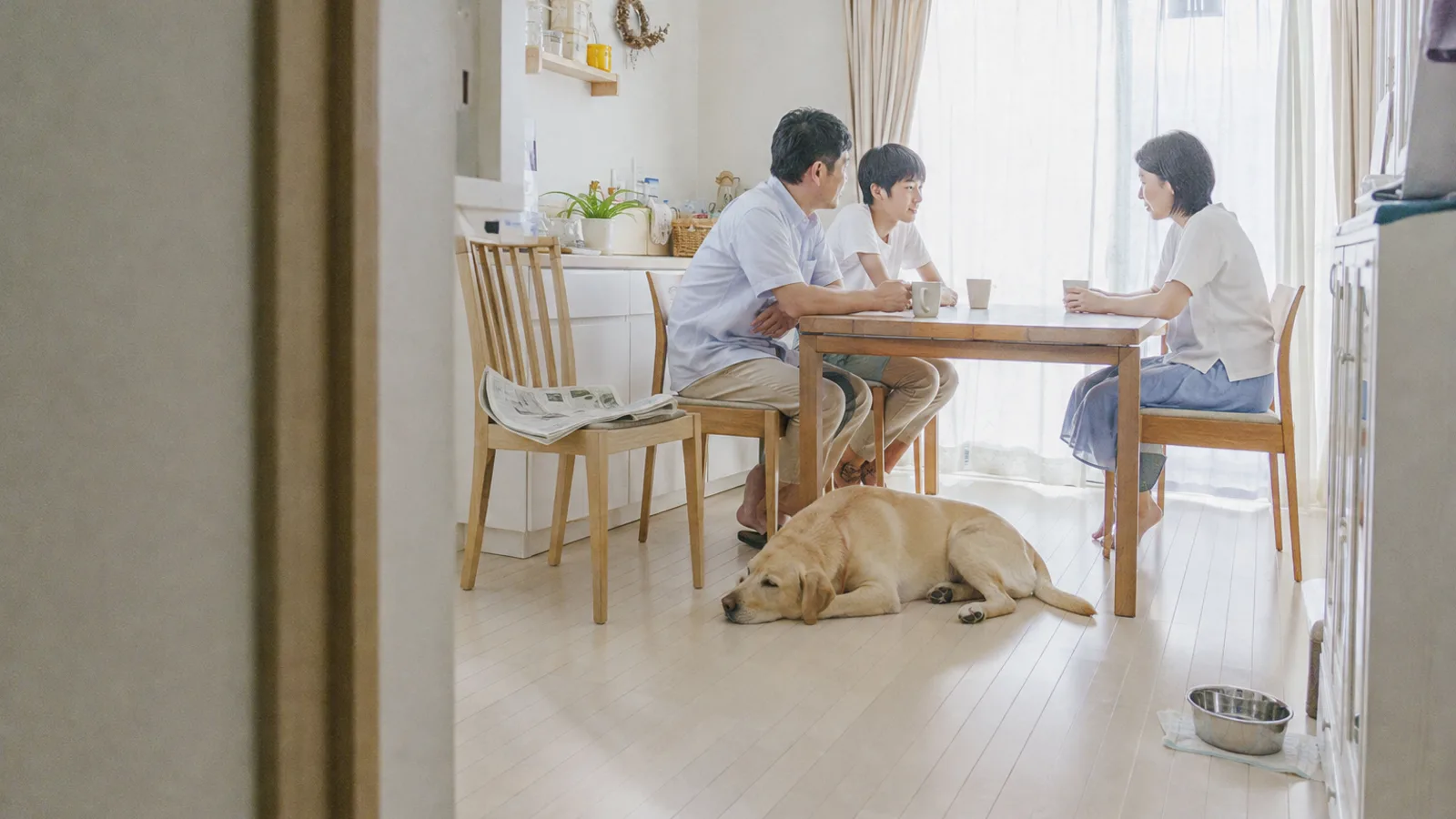 ダイニングテーブルでマグを前に静かに話す3人家族と、テーブルのそばの床に伏せるイエローのラブラドール・レトリーバー