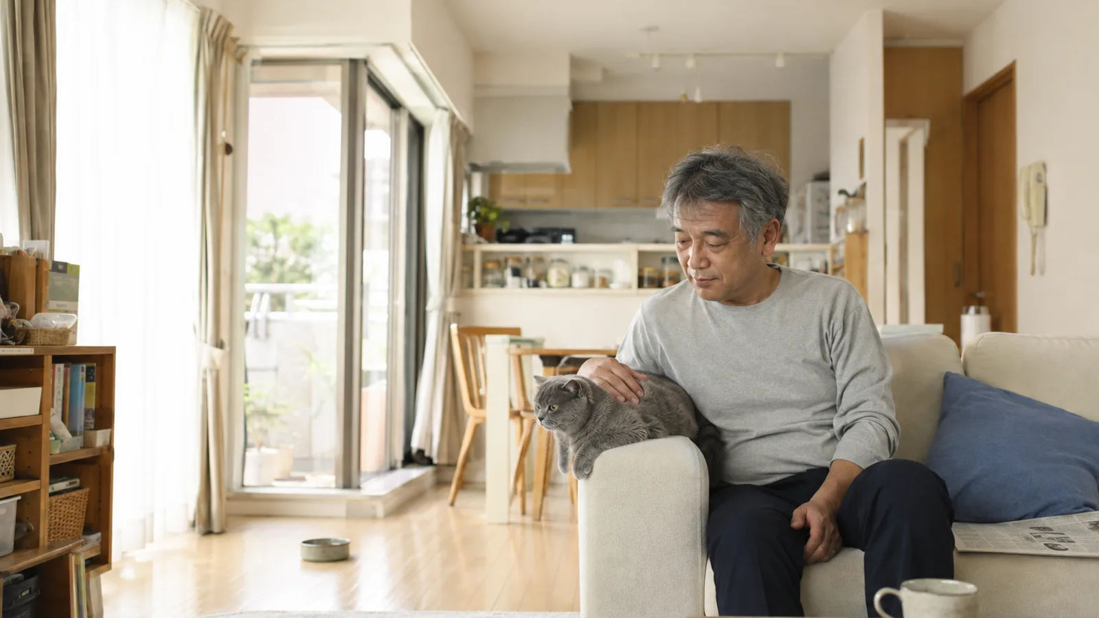 ソファの肘掛けに沿って寝そべる耳の小さな青灰色の猫と、背中に手を置いてくつろぐ男性