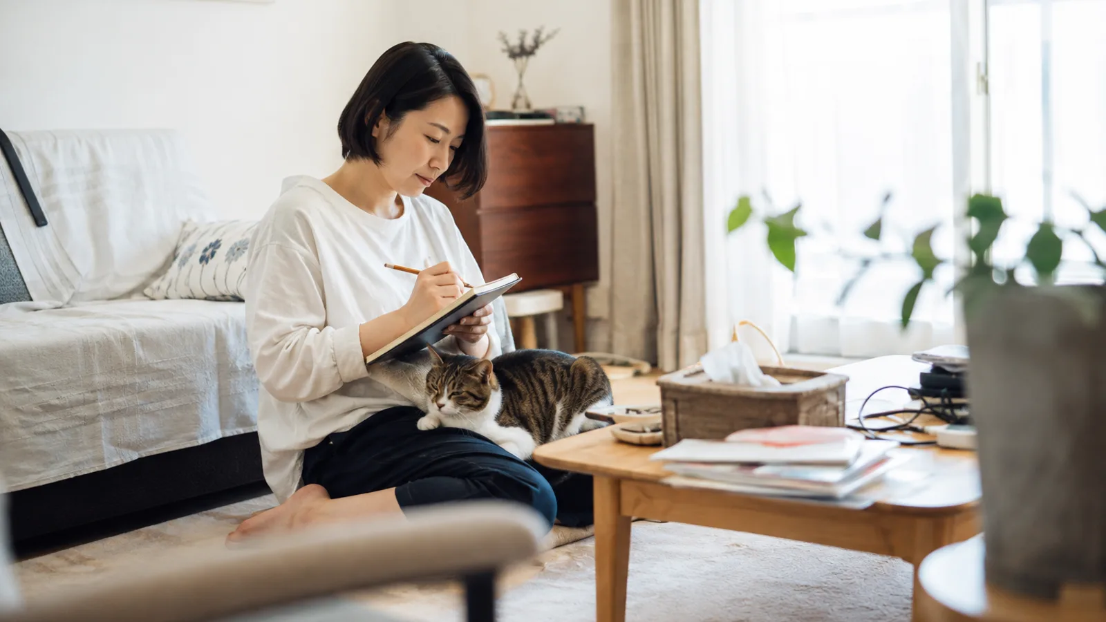 床に座った女性の膝に乗るキジトラ白の猫と、手に持った無地のノートに鉛筆で書いている女性