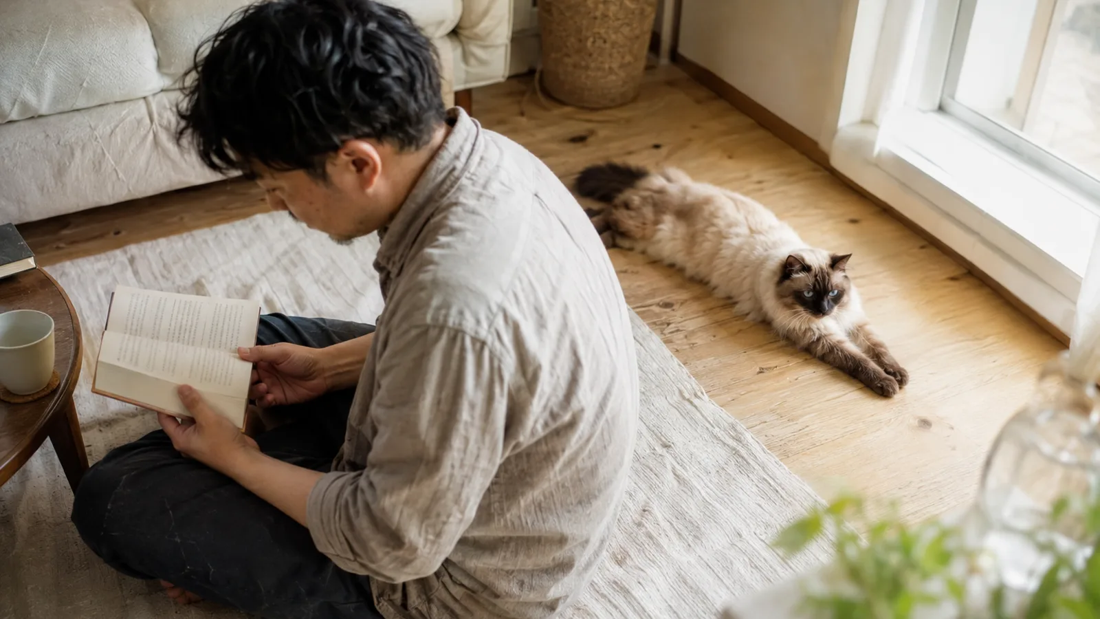 床で本を読む男性と、その少し後ろで前足を伸ばして寝そべる、青い目でクリーム色の体に濃い茶色のポイントがある長毛猫