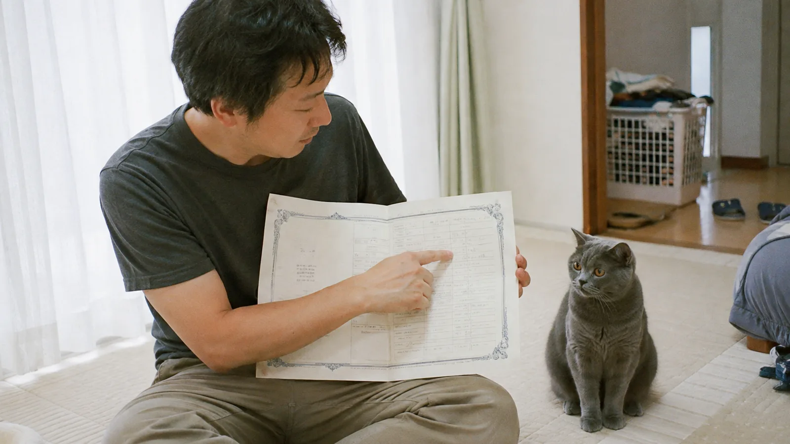 床で架空の猫の血統書を広げ、ご先祖の欄を指でなぞる男性と、隣に座る灰青色の短毛猫