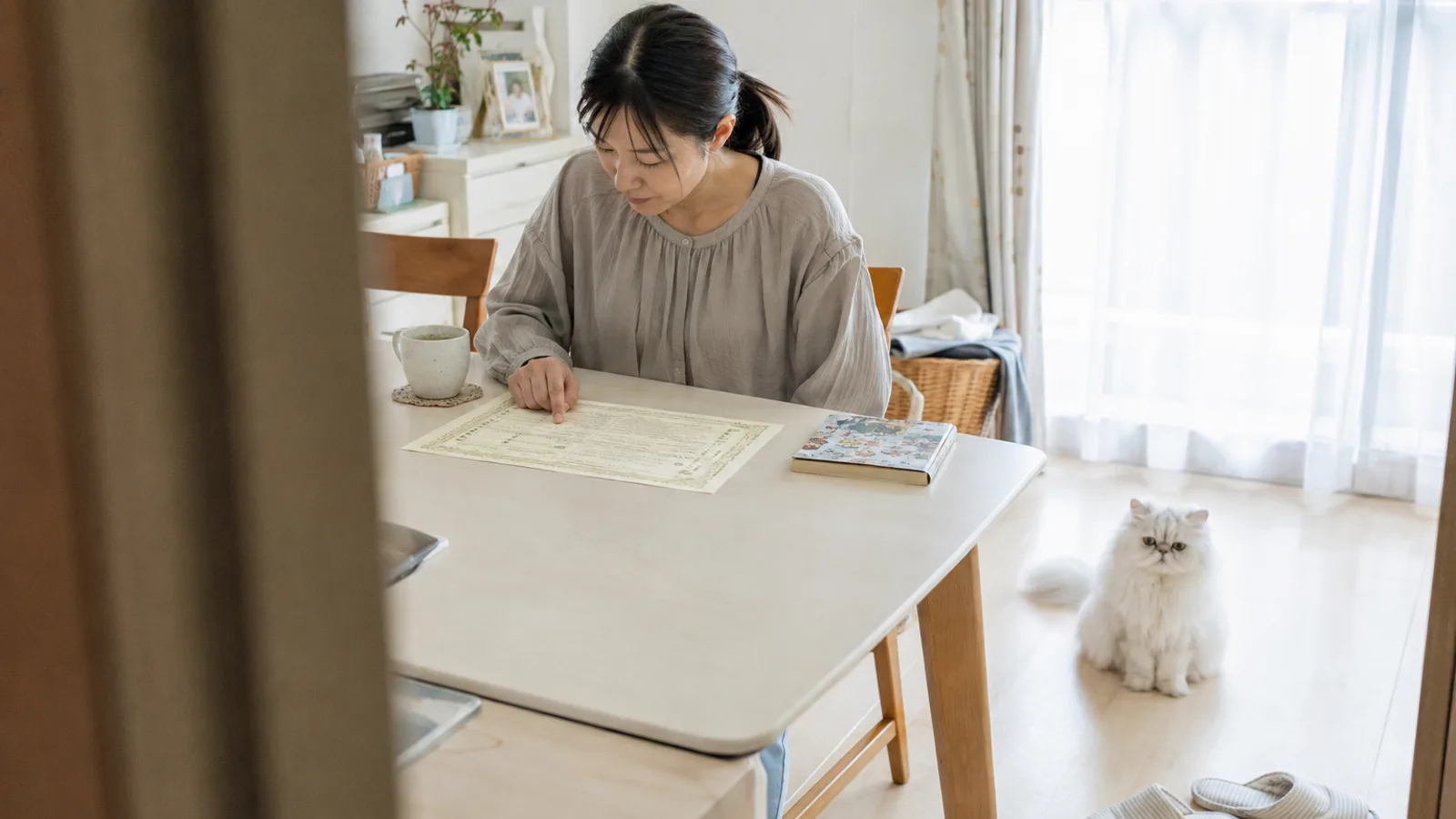 テーブルに平らに置いた架空の血統書を指でたどる女性と、少し離れた床に座る白い長毛の猫