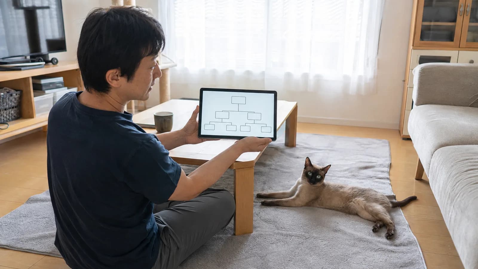 床でタブレットの家系図の画面を見る男性と、そばのラグに体を伸ばして伏せるトンキニーズ
