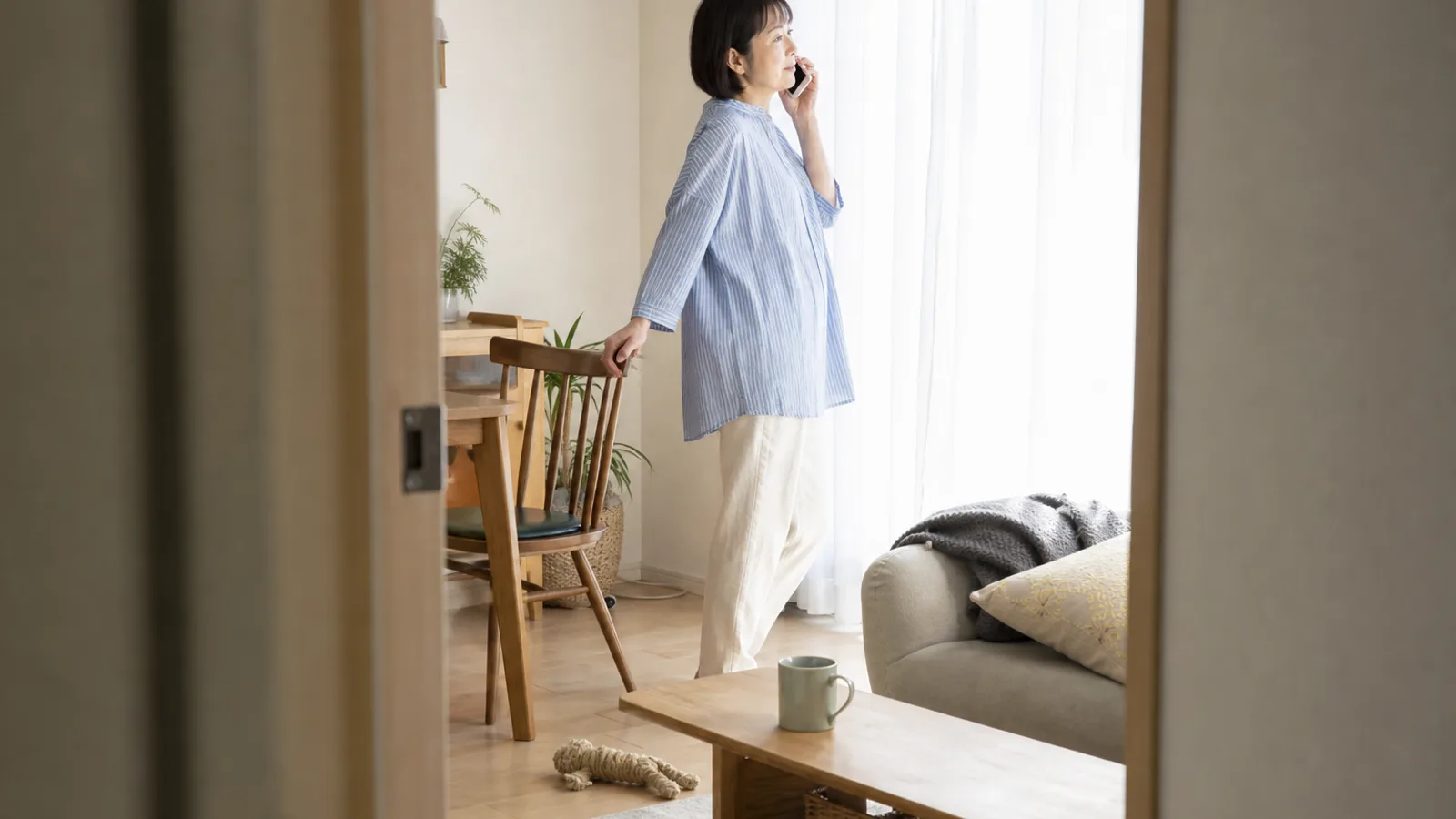 日の差す窓ぎわに立ち、スマホを耳に当てて静かに話を聞いている女性