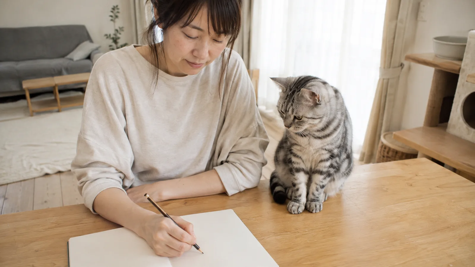 テーブルの無地のノートに鉛筆で書く女性と、すぐ横のテーブルに座って手元を見ているシルバータビーの猫